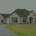 Front of Needville Cottage, a single-story stone home with a dark roof, a curved driveway and a green lawn.